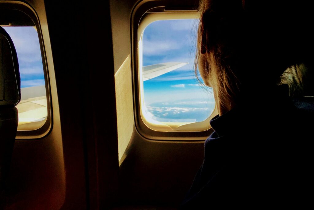 Silhouette of a woman looking out an airplane window at clouds and blue sky.